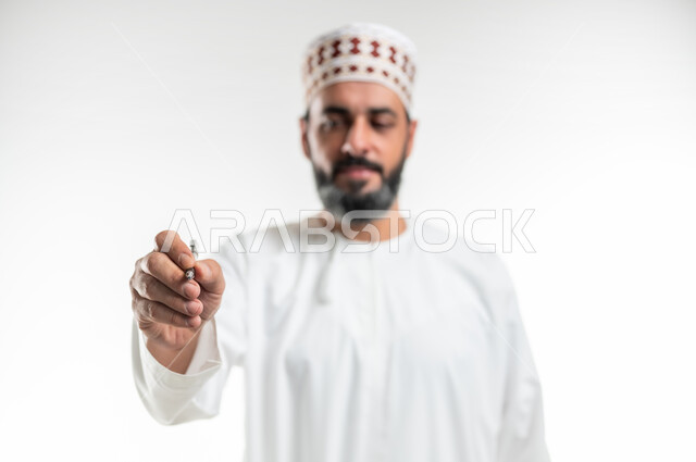الإستعداد للكتابة وتحديد شيء ما, مفهوم التعليم بالمناهج المقررة، تسجيل وتدوين الملاحظات، بورتريه مقرب لمعلم عربي خليجي عماني يرتدي الدشداشة و الكمه يمسك بيده قلم, إيماءات يدين تدل على الإمساك بعنصر ما، خلفية بيضاء
