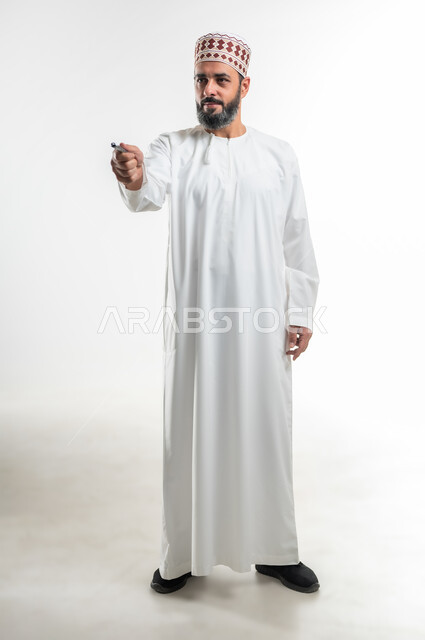 تعابير وحركات  يدين تدل على الاختيار والاشارة الى شخص، الإستعداد للكتابة وتحديد شيء ما, مفهوم التعليم بالمناهج المقررة، تسجيل وتدوين الملاحظات، بورتريه لمعلم عربي خليجي عماني يرتدي الدشداشة و الكمه يمسك بيده قلم, الطول الكامل للجسم، خلفية بيضاء