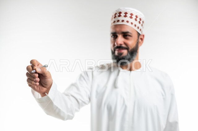 مفهوم التعليم بالمناهج المقررة، تسجيل وتدوين الملاحظات، الإستعداد للكتابة وتحديد شيء ما, بورتريه مقرب لمعلم عربي خليجي عماني يرتدي الدشداشة و الكمه يمسك بيده قلم, إيماءات يدين تدل على الإمساك بعنصر ما، خلفية بيضاء