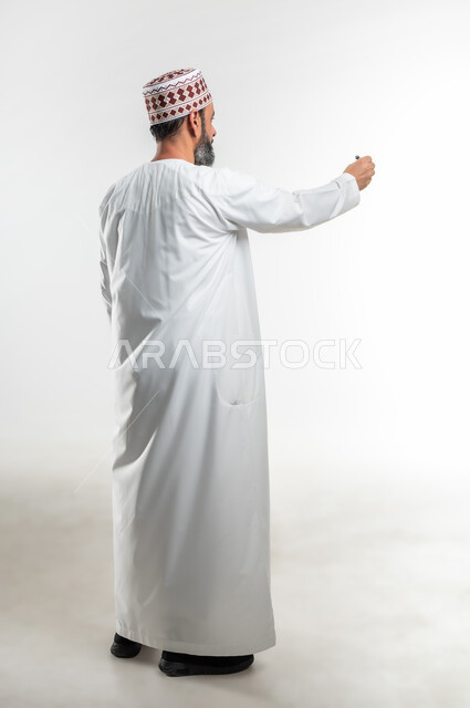 Preparing to write and define something, portrait from behind of an Arab Gulf Omani teacher wearing a dishdasha and a kummah holding a pen, hand gestures indicating holding an item, concept of education according to the prescribed curricula, full-length body image, white background