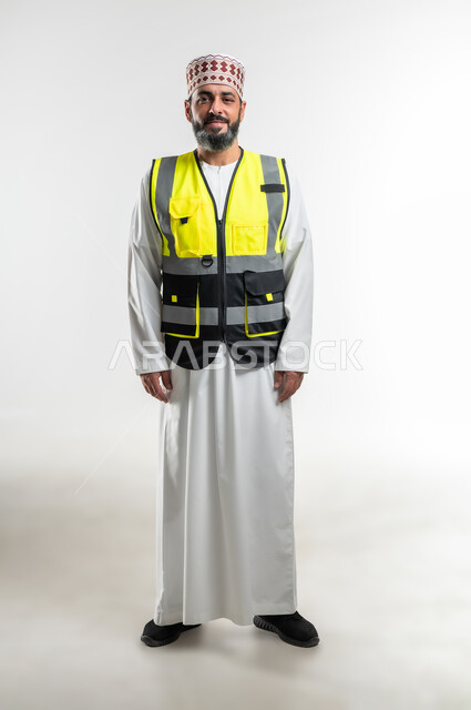 النظر الى الكاميرا والوقوف بإستقامة، تعابير السرور والسعادة والثقة بالنفس، العمل في القطاع الهندسي، مفهوم الهندسة والانشاءات المعمارية، بورتريه لمهندس عربي خليجي عماني مبتسم يرتدي الدشداشة و الكمه وسترة الحماية، الطول الكامل للجسم، خلفية بيضاء