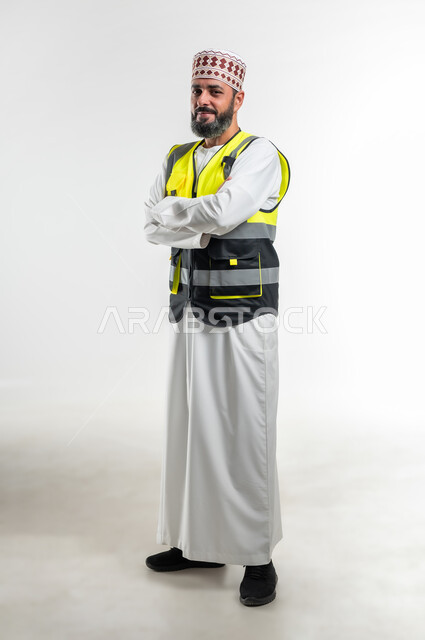 الوقوف بإستقامة و أيدي متقاطعه، تعابير السرور والثقة بالنفس، العمل في القطاع الهندسي، مفهوم الهندسة والانشاءات المعمارية، بورتريه لمهندس عربي خليجي عماني مبتسم يرتدي الدشداشة و الكمه وسترة الحماية ينظر إلى الكاميرا، الجسم بالطول الكامل، خلفية بيضاء