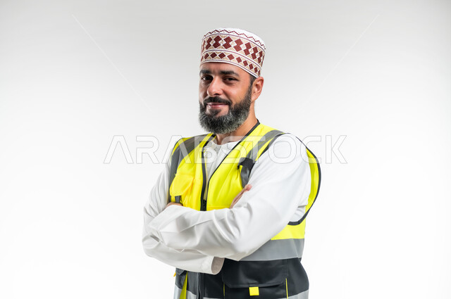 تعابير السرور والثقة بالنفس، الوقوف بإستقامة و أيدي متقاطعه، العمل في القطاع الهندسي، مفهوم الهندسة والانشاءات المعمارية، بورتريه مقرب لمهندس عربي خليجي عماني مبتسم يرتدي الدشداشة و الكمه وسترة الحماية ينظر إلى الكاميرا، خلفية بيضاء
