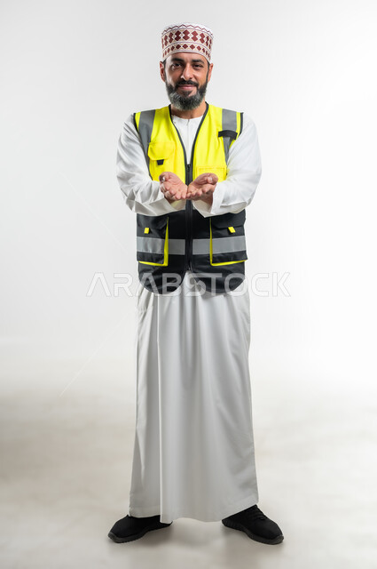 تعابير تدل على التقديم والعرض، العمل في القطاع الهندسي، مفهوم الهندسة والانشاءات المعمارية، بورتريه لمهندس عربي خليجي عماني يرتدي الدشداشة و الكمه وسترة الحماية ينظر الى الكاميرا و يفتح كفيه للأعلى بإيماءات حمل عنصر ما, صورة الجسم بالطول الكامل، خلفية بيضاء