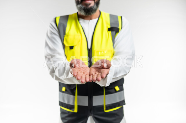 مفهوم الهندسة والانشاءات المعمارية، تعابير تدل على التقديم والعرض، العمل في القطاع الهندسي، بورتريه مقرب لمهندس عربي خليجي عماني يرتدي الدشداشة و الكمه وسترة الحماية يفتح كفيه للأعلى بإيماءات حمل عنصر ما, خلفية بيضاء