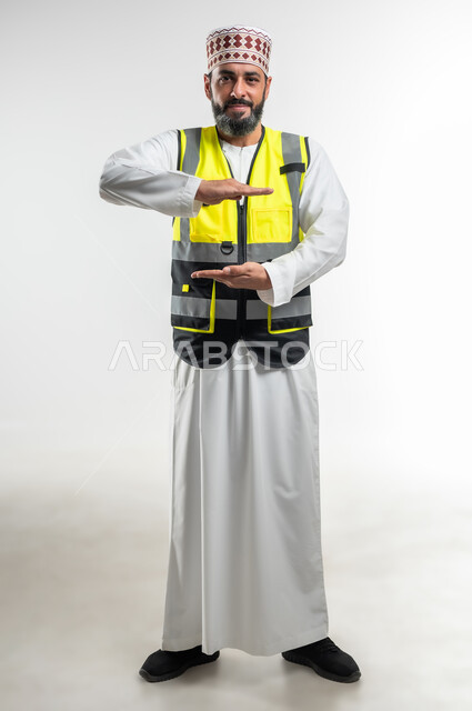 Holding and containing an element, working in the engineering sector, concept of engineering and architectural construction, portrait of an Arab Gulf Omani engineer wearing a dishdasha, a sleeve and a protective vest, opening his hands to display 3D holographic elements, full-length body, white background