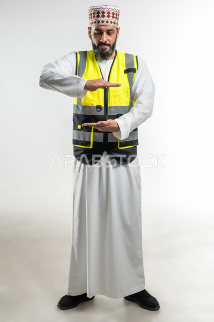 Holding and holding an element, working in the engineering sector, concept of engineering and architectural construction, portrait of an Arab Gulf Omani engineer wearing a dishdasha, a sleeve and a protective vest, opening his hands to display 3D holographic elements, full-length body, white background