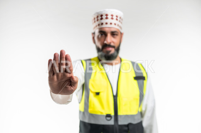 Stop and do not approach request, refusal, abstention and annoyance, concept of engineering and architectural construction, work in the engineering sector, close-up portrait of an Arab Gulf Omani engineer wearing a dishdasha, a sleeve and a protective vest, extending his hand forward with a raised palm in gestures of forbidding doing something, white background