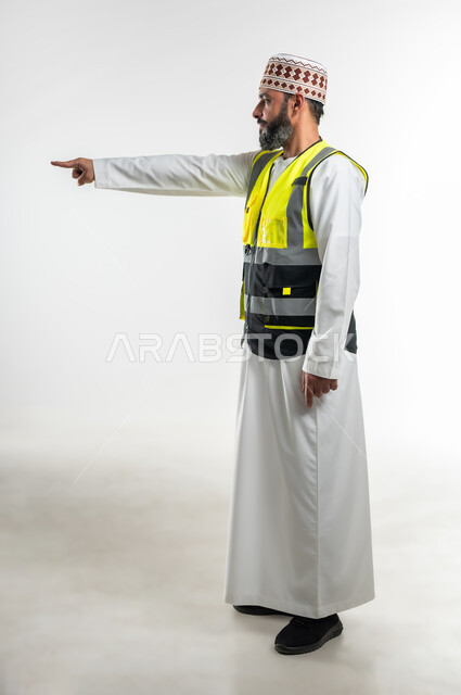 العمل في القطاع الهندسي، إشارات تدل على هذا المكان(هنا)، مفهوم الهندسة والانشاءات المعمارية، تحديد موقع معين، بورتريه من الجانب لمهندس عربي خليجي عماني يرتدي الدشداشة والكمه وسترة الحماية يرفع اصابعه السبابة ويشير الى شيء ما، الطول الكامل للجسم، خلفية بيضاء