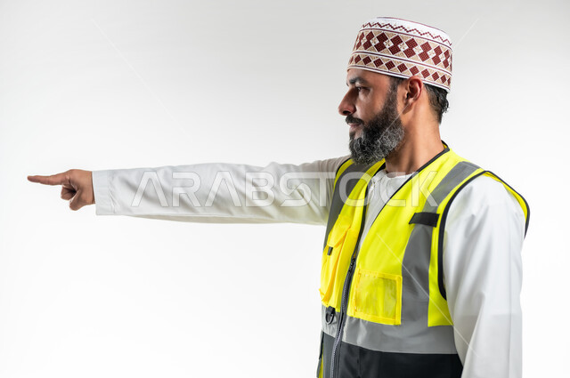 Concept of engineering and architectural construction, determining a specific location, working in the engineering sector, hand signals indicating this place (here), close-up portrait from the side of an Arab Gulf Omani engineer wearing a dishdasha, a sleeve and a protective vest, raising his index fingers and pointing to something, white background