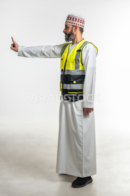 إشارات تدل على هذا المكان(هنا)، مفهوم الهندسة والانشاءات المعمارية، تحديد موقع معين، العمل في القطاع الهندسي، بورتريه من الجانب لمهندس عربي خليجي عماني يرتدي الدشداشة والكمه وسترة الحماية يرفع اصابعه السبابة ويشير الى شيء ما، الطول الكامل للجسم، خلفية بيضاء