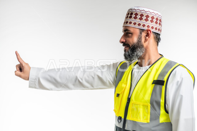 Determining a specific location, working in the engineering sector, the concept of engineering and architectural construction, hand signals indicating this place (here), close-up portrait from the side of an Arab Gulf Omani engineer wearing a dishdasha, a sleeve and a protective vest, raising his index fingers and pointing to something, white background