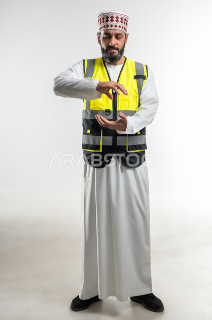 العمل في القطاع الهندسي، مفهوم الهندسة والانشاءات المعمارية، إحتواء وإمساك عنصر ما، بورتريه لمهندس عربي خليجي عماني يرتدي الدشداشة و الكمه وسترة الحماية يفتح يديه لعرض عناصرهولوجرامية ثلاثية الأبعاد الجسم بالطول الكامل، خلفية بيضاء