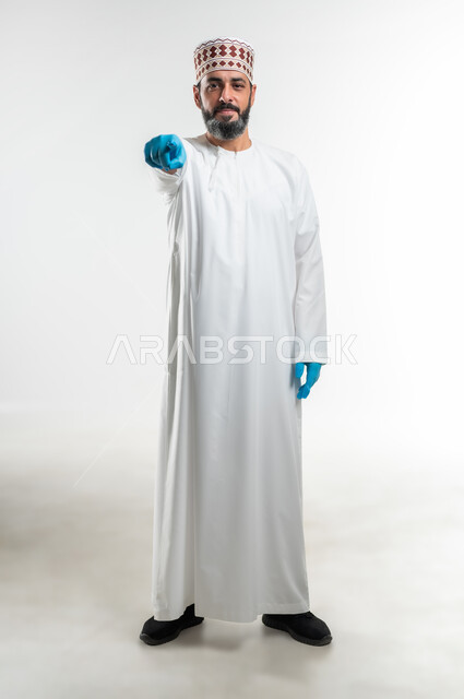 الشعور بالإنزعاج والرغبة بالإنتقام، إلقاء اللوم والتوبيخ والاتهام، طلب التوقف عن فعل الأخطاء, بورتريه لرجل عربي خليجي عماني يرتدي الدشداشة و الكمه وقفازات الحماية يرفع اصبع السبابة للاعلى لتحديد شخص ما ( أنت ), صورة الجسم بالطول الكامل, خلفية بيضاء