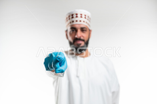 إلقاء اللوم والتوبيخ والاتهام، الشعور بالإنزعاج والرغبة بالإنتقام، طلب التوقف عن فعل الأخطاء, بورتريه مقرب لرجل عربي خليجي عماني يرتدي الدشداشة و الكمه وقفازات الحماية يرفع اصبع السبابة للاعلى لتحديد شخص ما ( أنت ), خلفية بيضاء