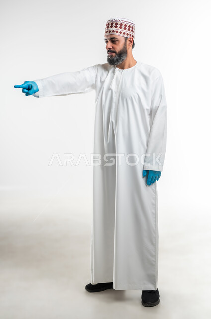 الاشارة إلى شيء ما، اشارات تحدد هذا المكان (هنا)، تعابير التحديد والتأكيد, بورتريه لرجل عربي خليجي عماني يرتدي الدشداشة والكمه وقفازات الوقاية يقف باستقامة و يشير بأصابع السبابة للاختيار، صورة الجسم بالطول الكامل، خلفية بيضاء