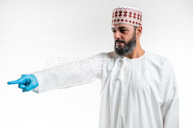 Gestures to indicate this place (here), pointing to something, expressions of specification and emphasis, close-up portrait of an Arab Gulf Omani man wearing a dishdasha, a sleeve and protective gloves, standing straight and pointing with index fingers to choose, white background