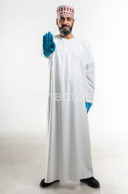 تعابير الحزم والجدية, مفهوم الرفض والامتناع والانزعاج, طلب التوقف وعدم الاقتراب, بورتريه لرجل عربي خليجي عماني يرتدي الدشداشة والكمه وقفازات الحماية يمد يده نحو الأمام مع رفع الكف بإيماءات النهي عن فعل امر ما, صورة الجسم بالطول الكامل، خلفية بيضاء