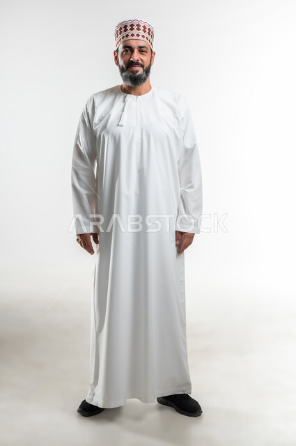 التقاط الصور الشخصية للوثائق الرسمية، الاهتمام بالأناقة والمظهر الخارجي, مفهوم الرجولة والثقة بالنفس، بورتريه لرجل عربي خليجي عماني مبتسم يرتدي الدشداشة و الكمه ينظر الى الكاميرا بإيماءات السرور والسعادة، صورة الجسم بالطول الكامل, خلفية بيضاء