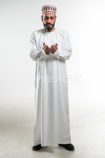 الإكثار من الدعاء في شهر رمضان المبارك، الإجتهاد في العبادة والحفاظ على الطاعات، بورتريه لرجل عربي خليجي عماني يرتدي الدشداشة و الكمه يرفع يديه ويدعو الله بإيماءات التوسل والتضرع، صورة الجسم بالطول الكامل, خلفية بيضاء
