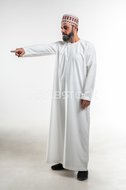 اشارات تحدد هذا المكان (هنا)، تعابير التحديد والتأكيد, الاشارة إلى شيء ما، بورتريه لرجل عربي خليجي عماني يرتدي الدشداشة والكمه يقف باستقامة و يشير بأصابع السبابة للاختيار، صورة الجسم بالطول الكامل، خلفية بيضاء