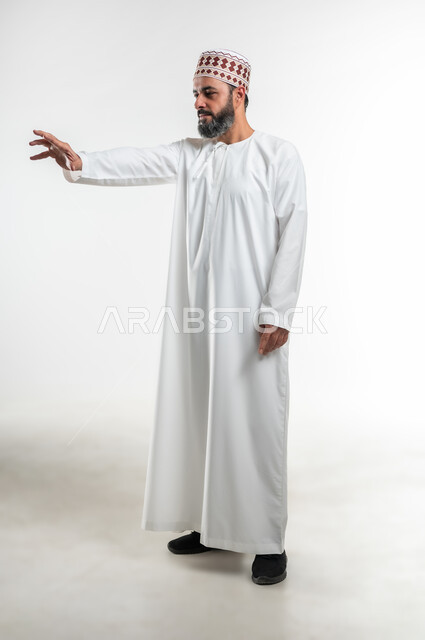 محاولة الامساك بشيء، طلب التوقف والامتناع عن فعل شيء ما، بورتريه لرجل عربي خليجي عماني يرتدي الدشداشة والكمه يقف باستقامة ويمد يده بتعابير تحدد هذا مكان (هنا)، ايماءات واشارات التحديد والتأكيد، الطول الكامل للجسم، خلفية بيضاء