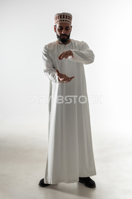 إمساك وإحتواء عنصر ما، بورتريه لرجل  عربي خليجي عماني يرتدي الدشداشة و الكمه يفتح يديه لعرض عناصر هولوجرامية ثلاثية الأبعاد, ايماءات وتعابير تدل على التركيز والدقة, صوره الجسم بالطول الكامل، خلفية بيضاء