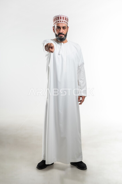 التوبيخ و الإتهام وإلقاء اللوم، طلب التوقف عن فعل الأخطاء، الشعور بالإنزعاج والرغبة بالإنتقام، بورتريه لرجل  عربي خليجي عماني يرتدي الدشداشة و الكمه يرفع اصبع السبابه الى الأمام لتحديد شخص ما ( أنت )، صورة الجسم بالطول الكامل، خلفية بيضاء