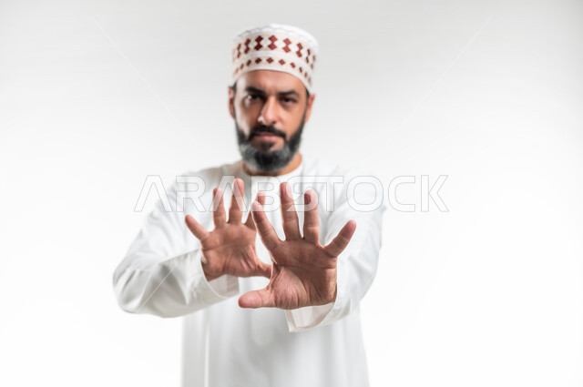 القلق والاحتياط والابتعاد عن شيء ما, حماية النفس من ضرر خارجي, بورتريه مقرب لرجل  عربي خليجي عماني يرتدي الدشداشة و الكمه يرفع يديه للاعلى بإيماءات التوقف وعدم الاقتراب، مفهوم الحذر وترك مسافة، خلفية بيضاء