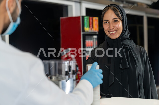 توفير وتجهيز الادوية اللازمة للمريضة،  إمرأة عربية خليجية إماراتية تشتري العقاقير الطبية من صيدلاني اماراتي يرتدي الكمامة الطبية للوقاية من فايروس كورونا، قراءة الروشتة الخاصة بالمريض، صرف الادوية الموجودة في الوصفات الطبية، بيع الادوية والمستلزمات الطبية