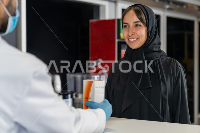 توفير وتجهيز الادوية اللازمة للمريضة،  إمرأة عربية خليجية إماراتية تشتري العقاقير الطبية من صيدلاني اماراتي يرتدي الكمامة الطبية للوقاية من فايروس كورونا، قراءة الروشتة الخاصة بالمريض، صرف الادوية الموجودة في الوصفات الطبية، بيع الادوية والمستلزمات الطبية