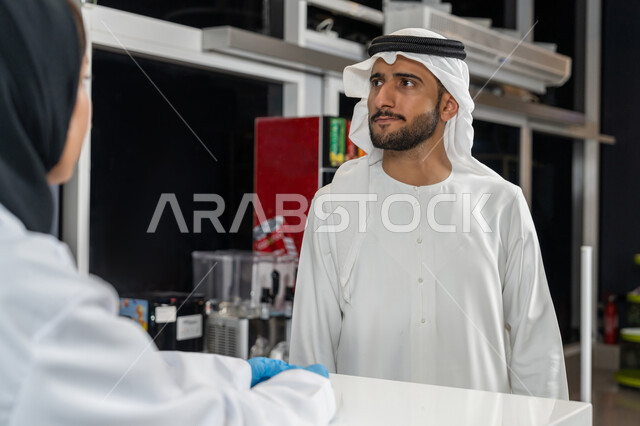 Benefiting from health services and care, describing the required medications and medical condition, an Arab Emirati Gulf man wearing a ghutra and a kandura asks the pharmacist for some medications from inside the pharmacy, selling medications and medical supplies