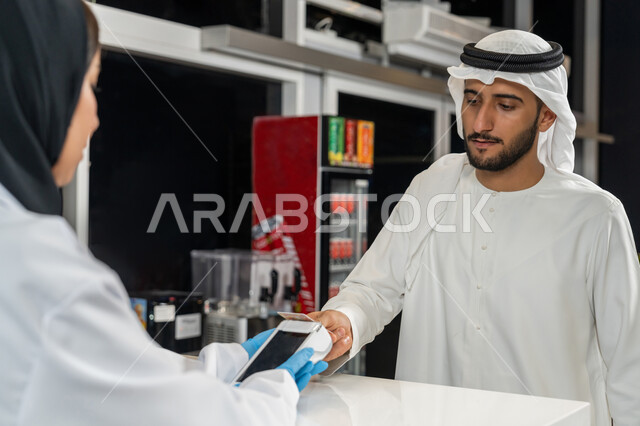 استخدام وسائل الدفع الالكتروني الحديثة والتكنولوجيا المتطورة، رجل عربي اماراتي خليجي يرتدي الغترة والكندورة يقوم بالدفع عن طريق الهاتف المحمول داخل الصيدلية، تسديد الفاتوره بالبطاقه البنكيه، بيع الادوية والمستلزمات الطبية، الاستفادة من الخدمات والرعاية الصحية