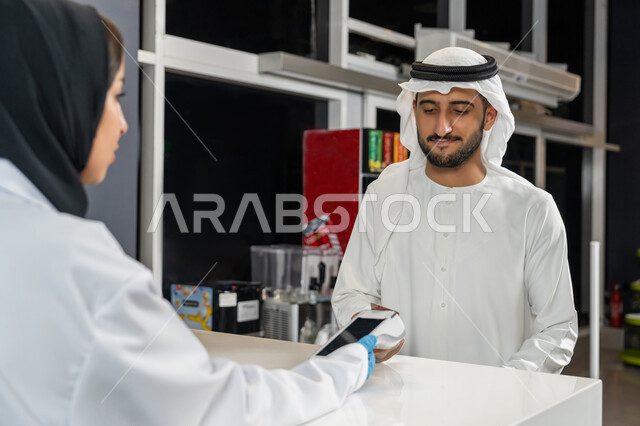 تسديد الفاتوره بالبطاقه البنكيه، استخدام وسائل الدفع الالكتروني الحديثة والتكنولوجيا المتطورة، رجل عربي اماراتي خليجي يرتدي الغترة والكندورة يقوم بالدفع عن طريق الهاتف المحمول داخل الصيدلية، بيع الادوية والمستلزمات الطبية، الاستفادة من الخدمات والرعاية الصحية