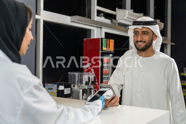 تسديد الفاتوره بالبطاقه البنكيه، استخدام وسائل الدفع الالكتروني الحديثة والتكنولوجيا المتطورة، رجل عربي اماراتي خليجي يرتدي الغترة والكندورة يقوم بالدفع عن طريق الهاتف المحمول داخل الصيدلية، بيع الادوية والمستلزمات الطبية، الاستفادة من الخدمات والرعاية الصحية