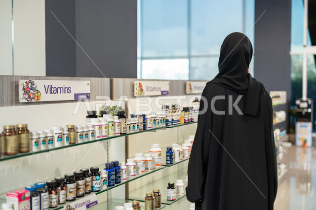 وصف الادوية المطلوبة والحالة المرضية، شراء الادوية والمستلزمات الطبية، الاستفادة من الخدمات الصحية، شابة سعودية تقوم باختيار المنتجات الطبية، صورة من الخلف لإمراة عربية خليجية اماراتية محجبة ترتدي العباءة السوداء داخل الصيدلية