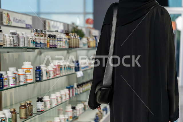 وصف الادوية المطلوبة والحالة المرضية، شراء الادوية والمستلزمات الطبية، الاستفادة من الخدمات الصحية، شابة سعودية تقوم باختيار المنتجات الطبية، صورة من الخلف لإمراة عربية خليجية اماراتية محجبة ترتدي العباءة السوداء داخل الصيدلية