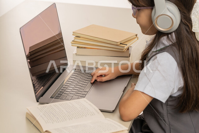 A diligent student sitting at a study table, the concept of e-learning, distance learning using modern technology, a portrait of an Arab Gulf Emirati student wearing a school uniform and headphones following lessons using a computer, white background