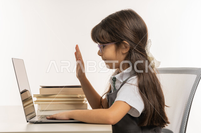 طالبة مجتهدة تجلس على طاولة الدارسة ترفع يدها للإجابة، المشاركة الفعالة للطلاب ومتابعة الدروس والواجبات من خلال منصات التعلم عن بعد، بورتريه لتلميذة عربية خليجية اماراتية ترتدي الزي المدرسي تحضر دروسها عن طريق جهاز اللابتوب، خلفية بيضاء