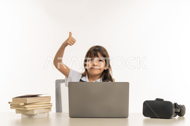 ايماءات وجه ويدين تدل على والقبول والموافقة، التعلم عن بعد باستخدام جهاز الحاسب المحمول، بورتريه لطالبة عربية خليجية اماراتية ترتدي الزي المدرسي و بجانبها نظارات الواقع الافتراضي ترفع اصبع الابهام للأعلى، تلميذة مجتهدة مبتسمة تنظر إلى الكاميرا، خلفية بيضاء