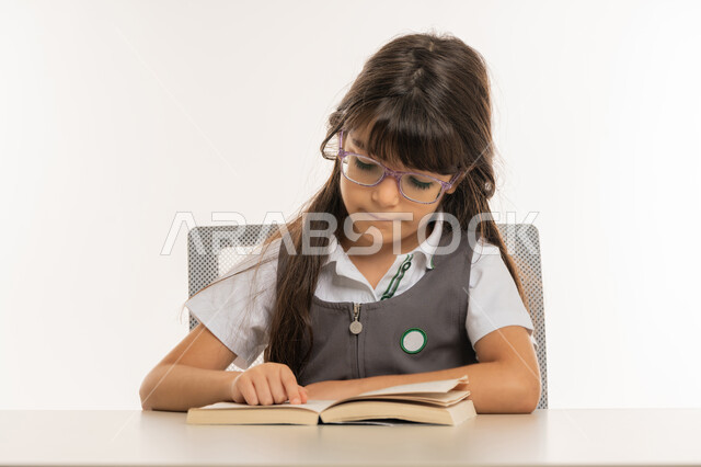 تلميذة مجتهدة تقوم بتدوين المعلومات لتمكين وفهم الدروس, اكتساب المعرفة من كتابة الواجبات، القيام بالمهام التعليمية الفعالة, بورتريه مقرب لطالبة عربية خليجية إماراتية ترتدي المريول المدرسي وتجلس على طاولة الدراسة, ايماءات الاندماج والتركيز، خلفية بيضاء