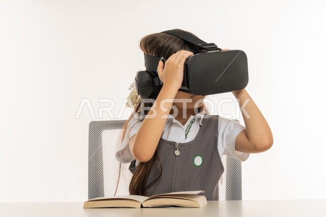 اكاديميات ومدارس متطورة، اتباع طرق تعليمية جديدة، بورتريه لتلميذة عربية خليجية اماراتية ترتدي المريول المدرسي تمسك بيديها نظارة العالم الافتراضي، طالبة مجتهدة تجلس على طاولة الدراسة وبجانبها الكتب، جمع التعلم مع الترفيه بواسطة التقنية الحديثة، خلفية بيضاء