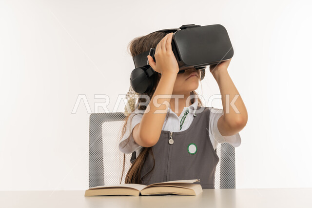 Combining learning with entertainment using modern technology, advanced academies and schools, following new educational methods, portrait of an Arab Gulf Emirati student wearing a school uniform holding virtual world glasses in her hands, a diligent student sitting at a study table with books next to her, white background