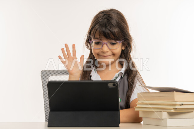 المشاركة الفعالة للطلاب ومتابعة الدروس والواجبات من خلال منصات التعلم عن بعد، بورتريه لتلميذة عربية خليجية اماراتية ترتدي الزي المدرسي تحضر دروسها عن طريق جهاز التابلت الإلكتروني، طالبة مجتهدة تجلس على طاولة الدارسة ترفع يدها للإجابة، خلفية بيضاء