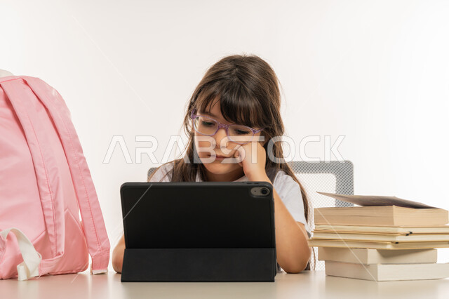 مفهوم التعليم الإلكتروني، التعليم عن بعد باستخدام التكنولوجيا الحديثة، طرق تعلمية جديدة ومتطورة، بورتريه لطالبة عربية خليجية اماراتية ترتدي المريول المدرسي تقوم بمتابعة الدروس باستخدام جهاز التابلت التقني، تلميذة مجتهدة تجلس على طاولة الدراسة، خلفية بيضاء