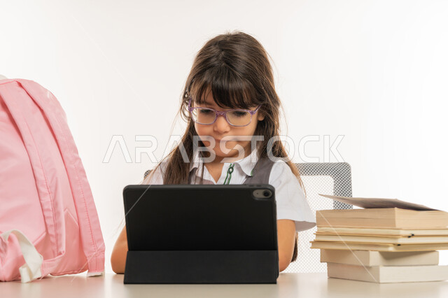 طرق تعلمية جديدة ومتطورة، بورتريه لطالبة عربية خليجية اماراتية ترتدي المريول المدرسي تقوم بمتابعة الدروس باستخدام جهاز التابلت التقني، تلميذة مجتهدة تجلس على طاولة الدراسة، مفهوم التعليم الإلكتروني، التعليم عن بعد باستخدام التكنولوجيا الحديثة، خلفية بيضاء