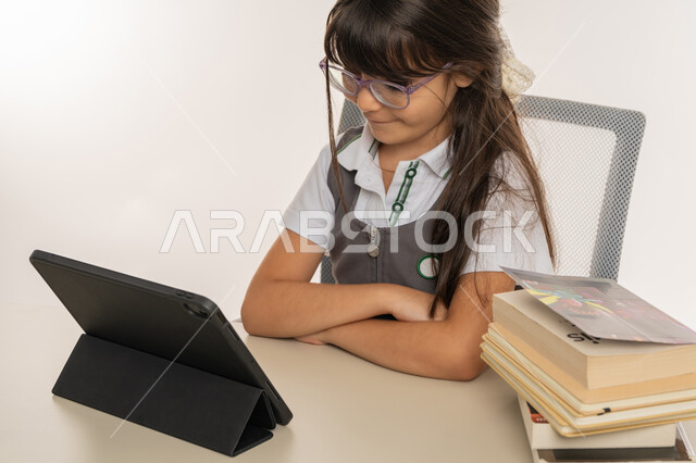 التعليم عن بعد باستخدام التكنولوجيا الحديثة، طرق تعلمية جديدة ومتطورة، بورتريه لطالبة عربية خليجية اماراتية ترتدي المريول المدرسي تقوم بمتابعة الدروس باستخدام جهاز التابلت التقني، تلميذة مجتهدة تجلس على طاولة الدراسة، مفهوم التعليم الإلكتروني، خلفية بيضاء