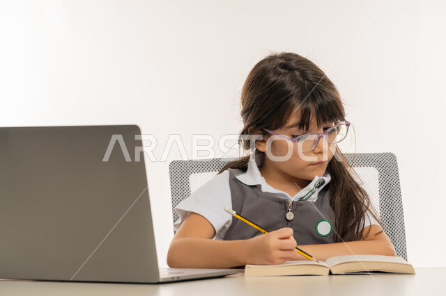 المذاكرة و الدراسة عن بعد، مفهوم التعليم الإلكتروني، طالبة مجتهدة تراجع دروسها من الكتب وتستخدم الحاسوب المحمول، متابعة الحصص و حل الواجبات عبر الانترنت، بورتريه مقرب لتلميذة خليجية عربية إماراتية ترتدي اليونيفورم المدرسي و تدرس في المنزل، خلفية بيضاء