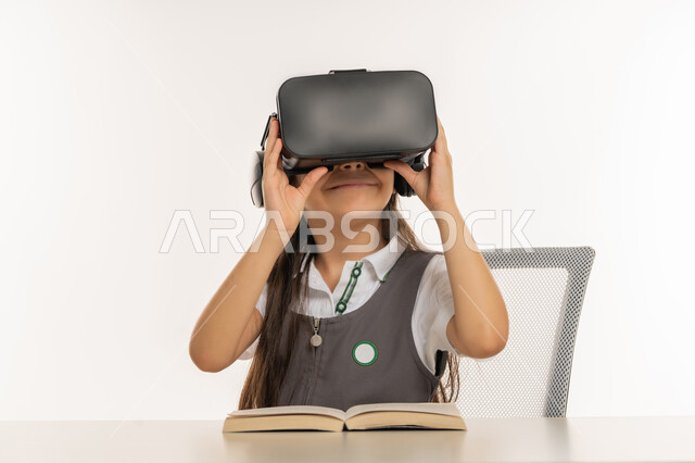 طالبة مجتهدة تجلس على طاولة الدراسة وبجانبها الكتب، جمع التعلم مع الترفيه بواسطة التقنية الحديثة، اكاديميات ومدارس متطورة، اتباع طرق تعليمية جديدة، بورتريه لتلميذة عربية خليجية اماراتية ترتدي المريول المدرسي تمسك بيديها نظارة العالم الافتراضي، خلفية بيضاء