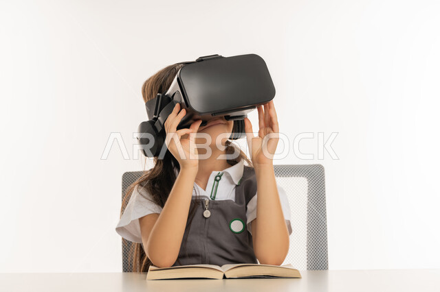 اتباع طرق تعليمية جديدة، بورتريه لتلميذة عربية خليجية اماراتية ترتدي المريول المدرسي تمسك بيديها نظارة العالم الافتراضي، طالبة مجتهدة تجلس على طاولة الدراسة وبجانبها الكتب، جمع التعلم مع الترفيه بواسطة التقنية الحديثة، اكاديميات ومدارس متطورة، خلفية بيضاء